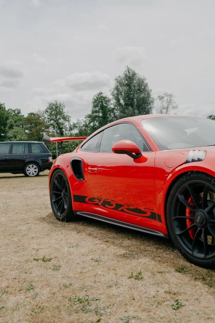 A red Porsche 911 GT3 RS sports car parked on grass with a black station wagon in the background, trees, and a cloudy sky.