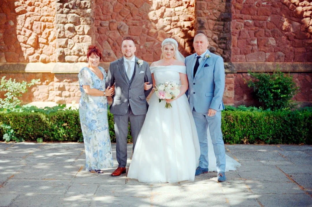 A wedding party with three adults and a bride in a white dress, standing outdoors in front of a brick wall and greenery.