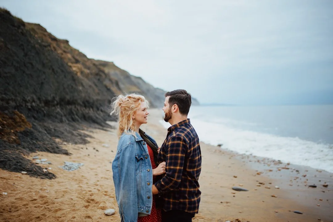 A young couple standing close on a beach, facing each other and smiling, with cliffs and the ocean in the background.