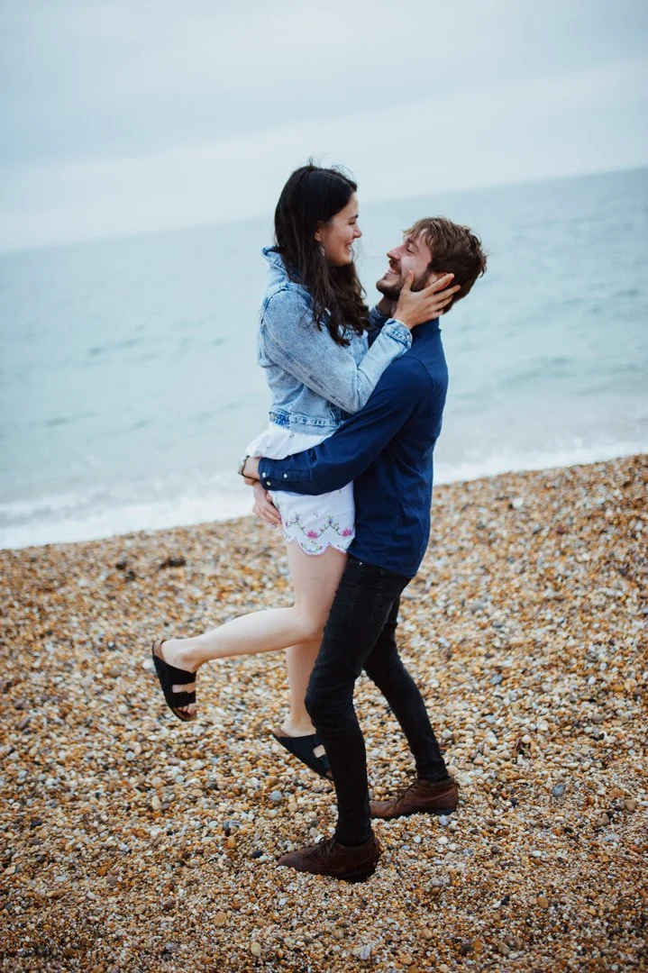 A man and woman on a pebble beach, with the woman being lifted by the man. They are smiling at each other, with the ocean in the background.