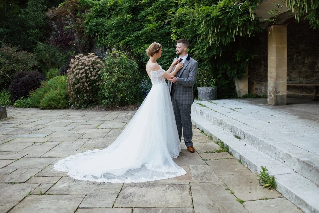 A bride and groom standing outdoors, holding hands and smiling at each other, surrounded by greenery, during their wedding.