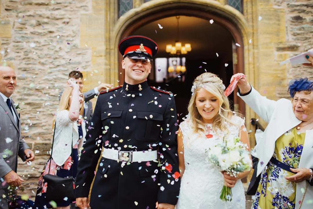 man waring military uniform walking with bride smiling and laughing, there is confetti being thrown behind them