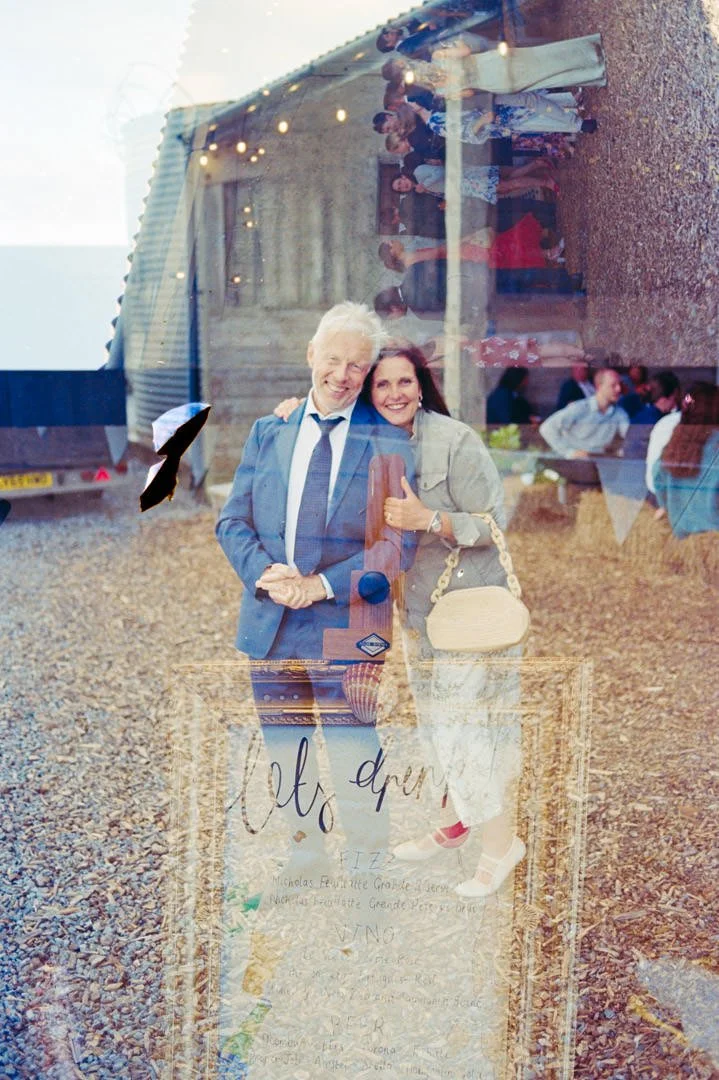 A reflection of a smiling couple taking a selfie outside a rustic building during an event, with other people visible in the background through the reflection.