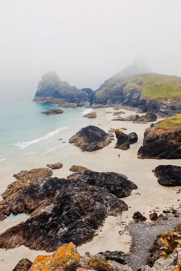 A foggy coastal scene with large rocks on a sandy beach, green cliffs, and mist-covered mountains in the background.