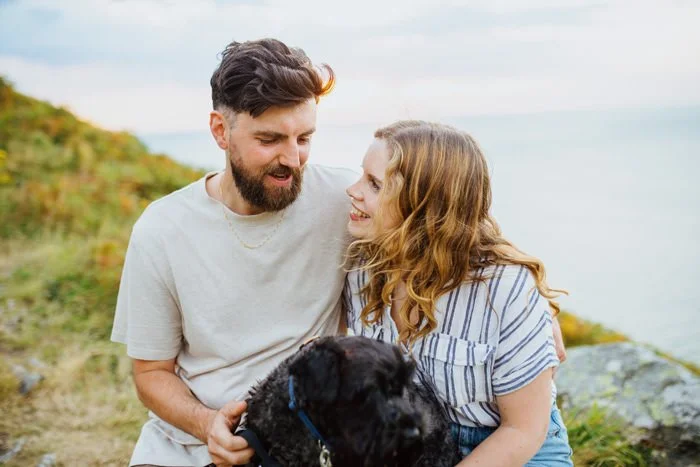 A young couple with a black dog outdoors near water and grassy hills.