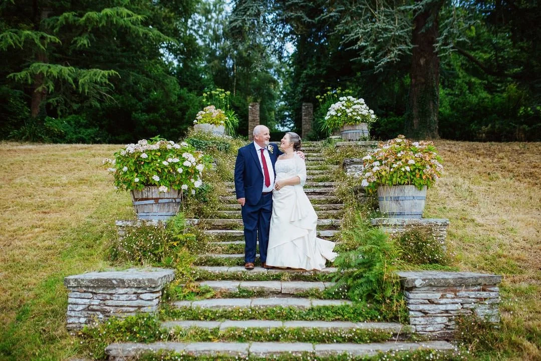 bride and groom on steps in floral garden under large trees, Hestercombe House