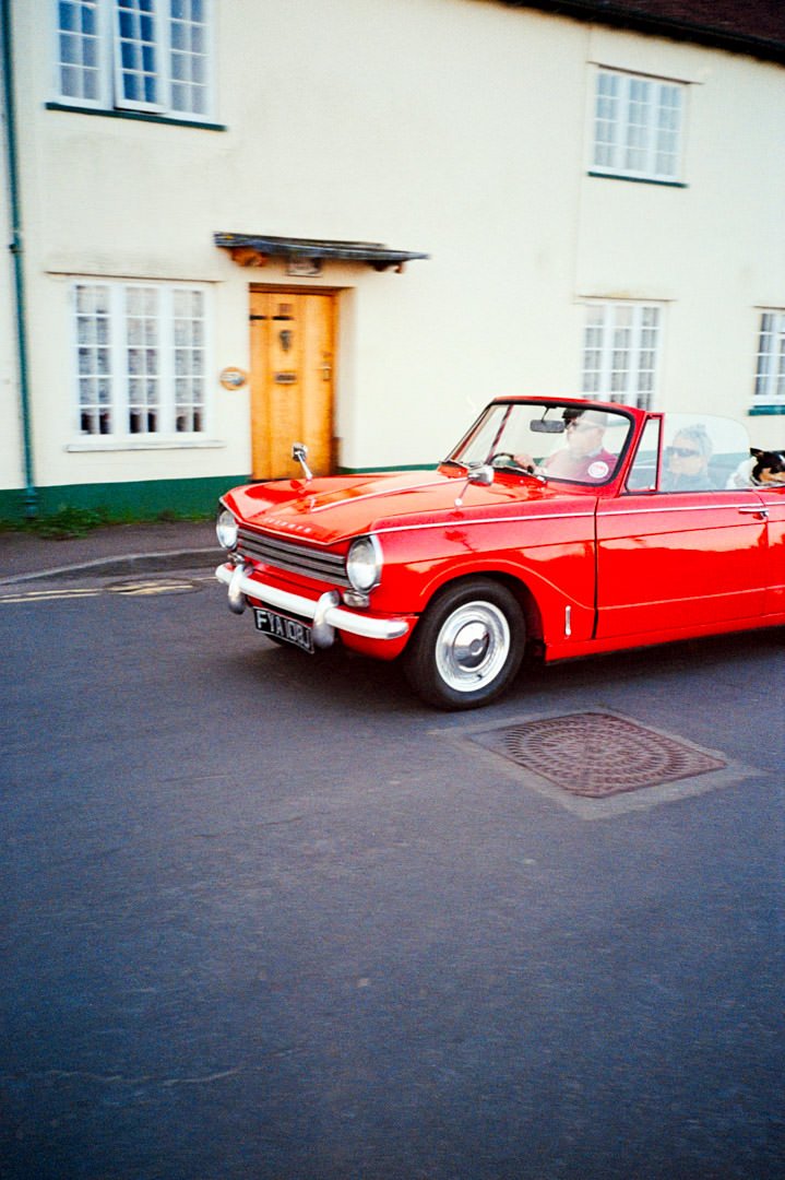 red classic convertible car driving with cottage houses in the background