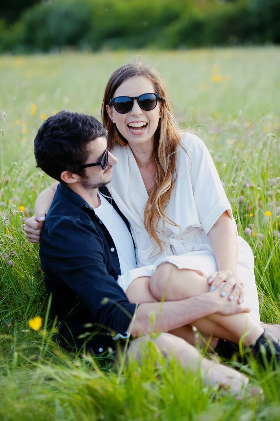 A man and a woman sitting together in a grassy field, laughing and enjoying each other's company. Both are wearing sunglasses, and the woman has long red hair while the man has dark hair and beard.