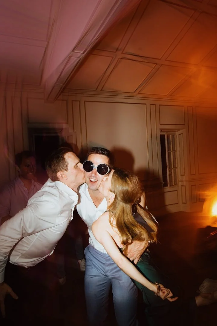 Three people at a party, two men and a woman, are sharing a kiss on the cheeks while smiling, with one of the men wearing sunglasses. The background shows a room with paneling, dim lighting, and a window, creating a lively, festive atmosphere.