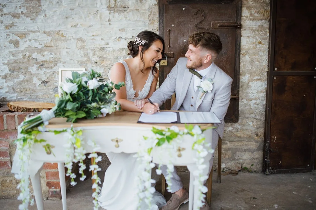 bride and groom sat at table singing a large book. they are smiling and looking at each other