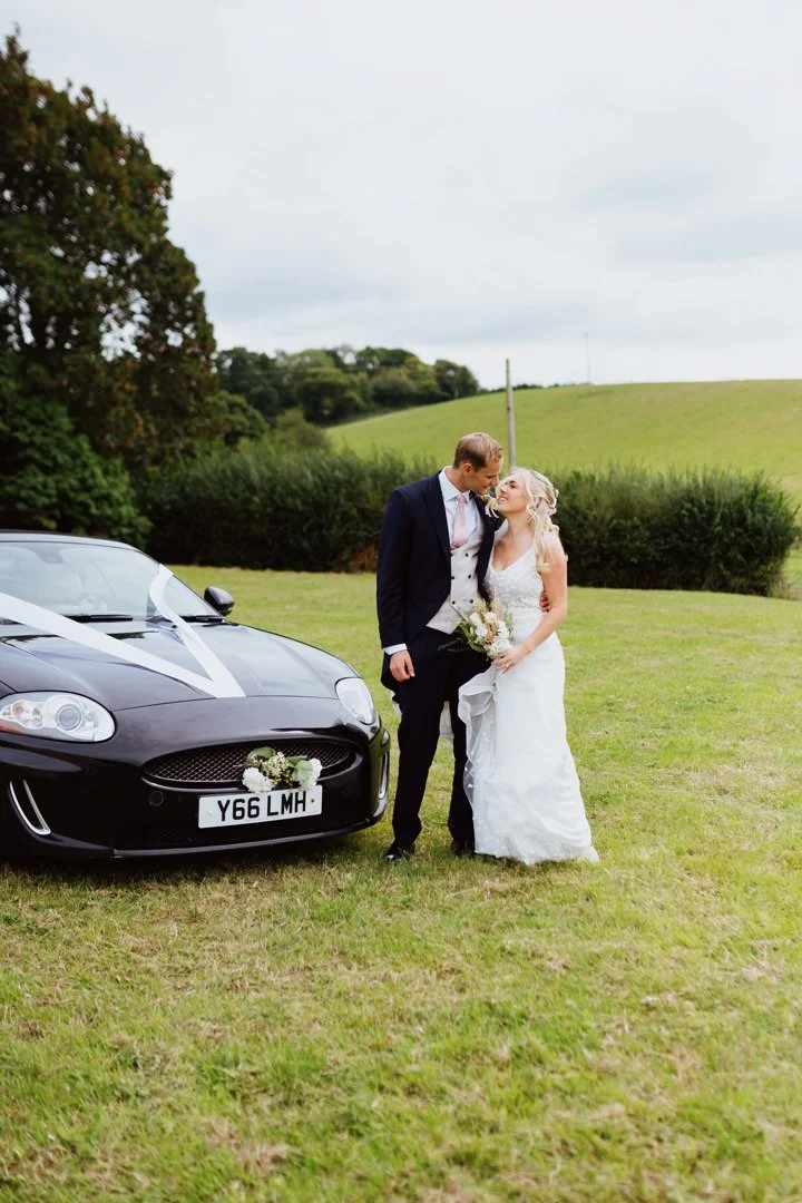 A newlywed couple standing on a grassy field beside a black wedding car decorated with white ribbons and flowers, with green hills and trees in the background.