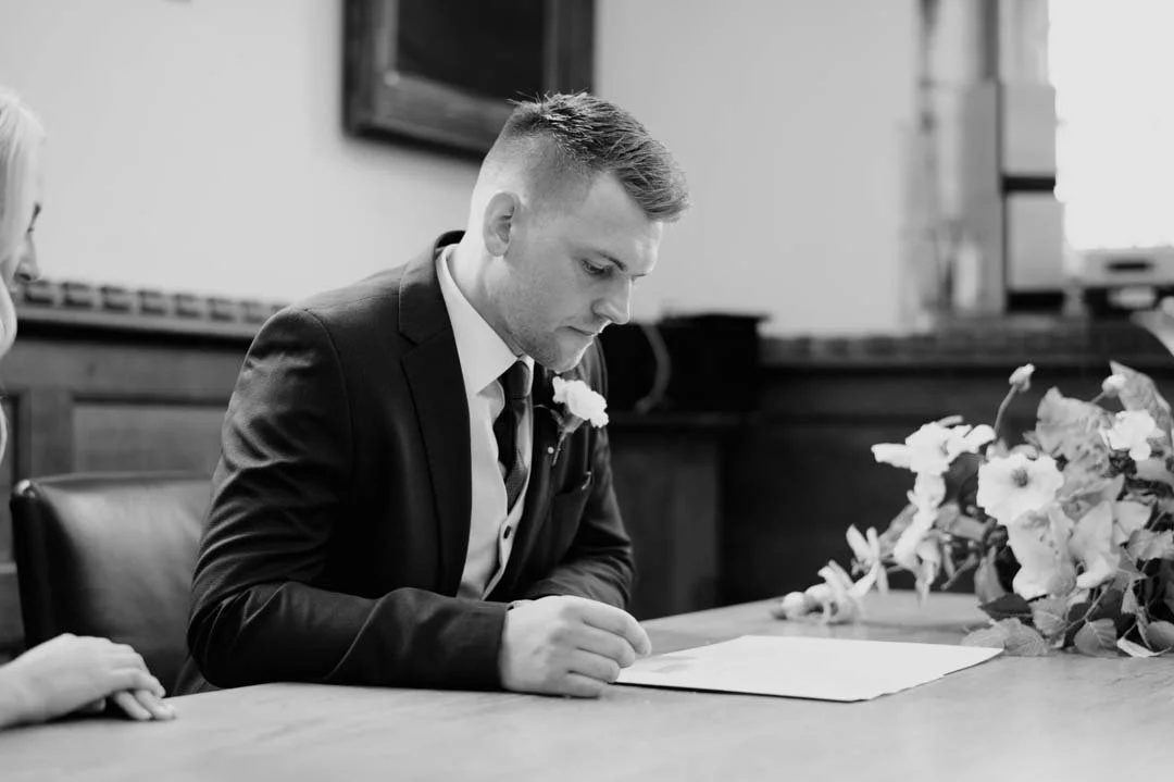 man waring blue suit signing paperwork, there are large wedding flowers on the table