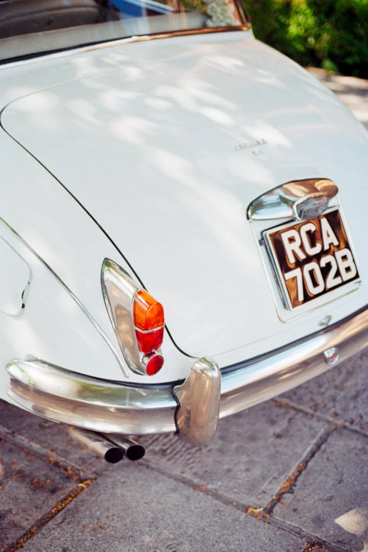 Rear view of a vintage white car parked on the street, showing the car's tail lights, chrome bumper, and a license plate with the registration number 'RCA 702B'.