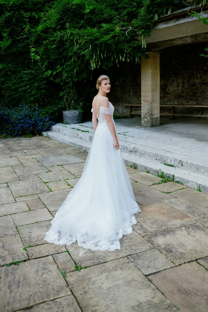 A woman in a white wedding dress standing outdoors on a stone patio, with greenery and a concrete structure in the background.