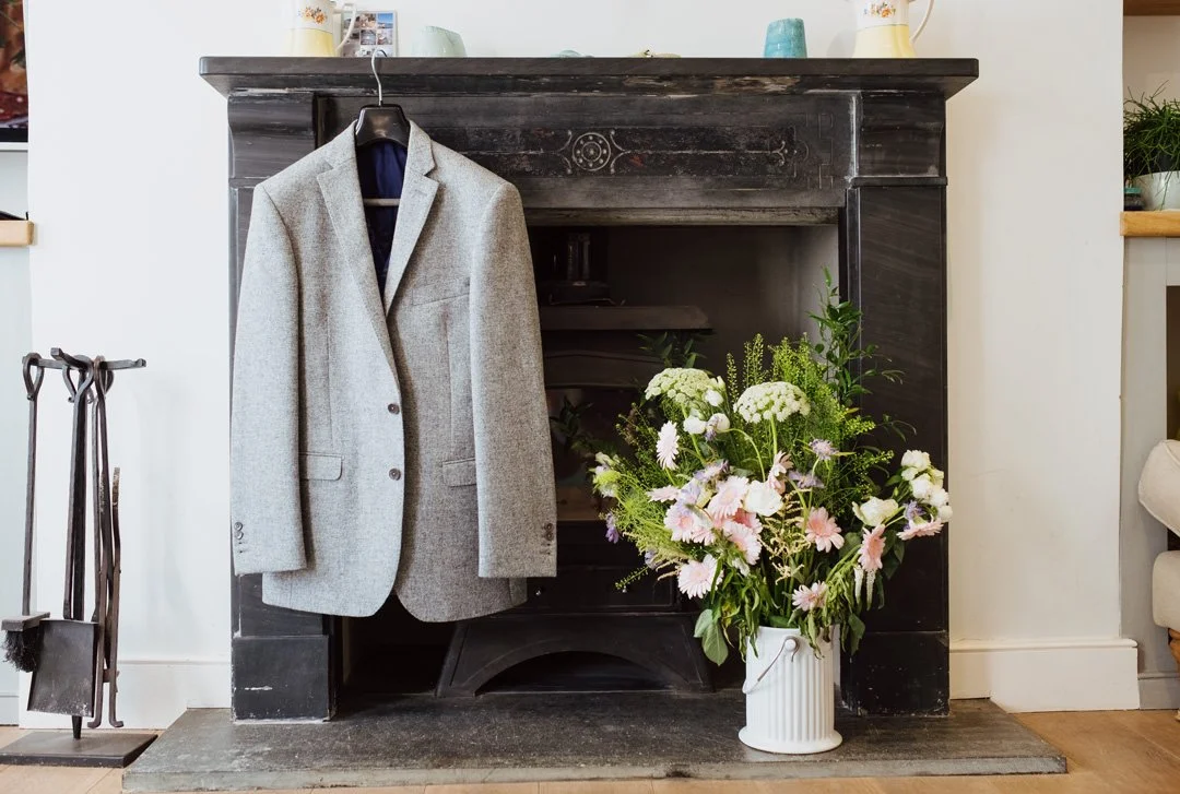 Grey wool blazer hanging on a black fireplace mantel next to a large bouquet of white and light pink flowers in a white vase on the floor.