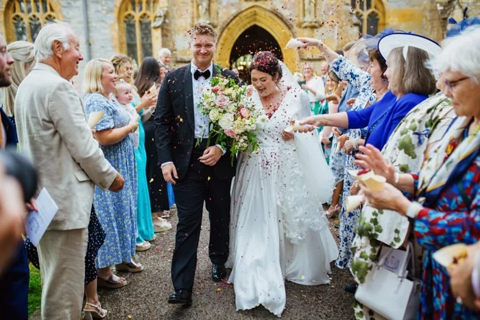 A bride and groom walking through a crowd of friends and family outside a church, celebrating their wedding with confetti and smiles.