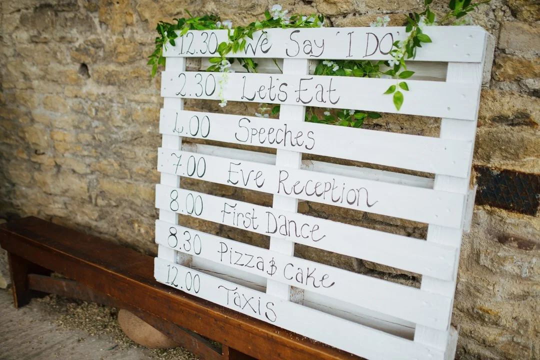 wooden pallet with writing showing timings for a wedding. there are green plants hanging over the front
