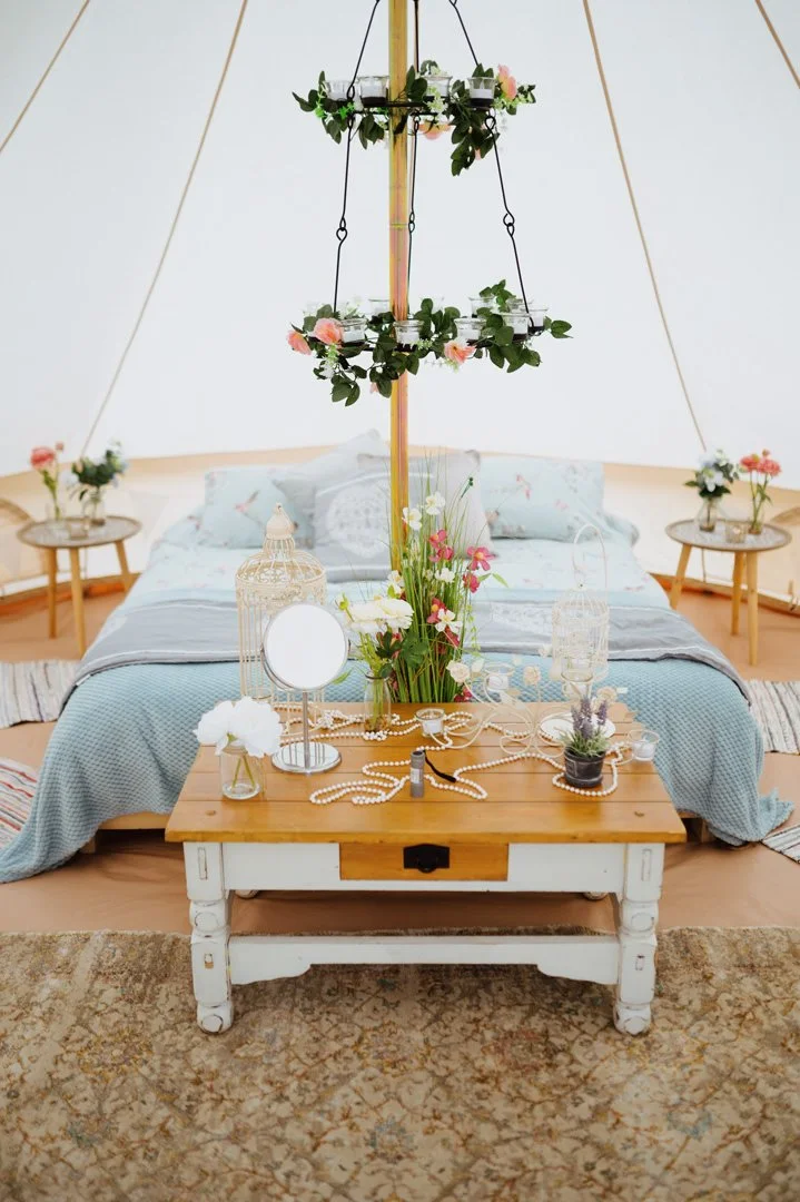 Interior of a cozy bedroom in a tipi with a bed, floral decorations, and a rustic wooden table with decorative items.