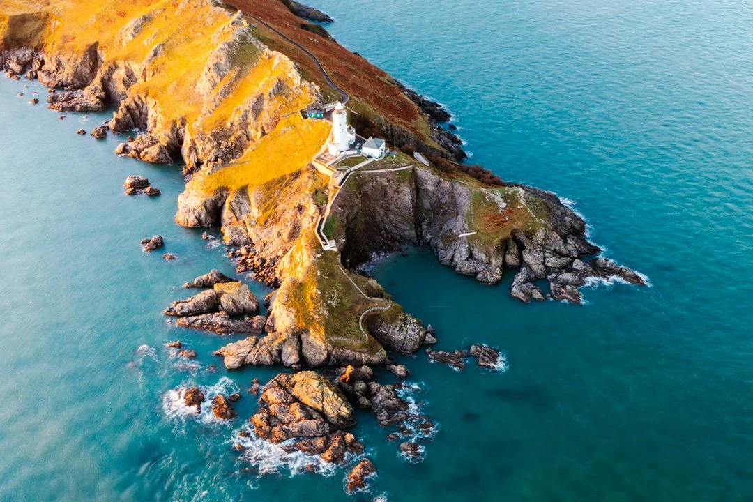 Aerial view of a rocky coastal area with cliffs, a lighthouse, and a small building, surrounded by the ocean.