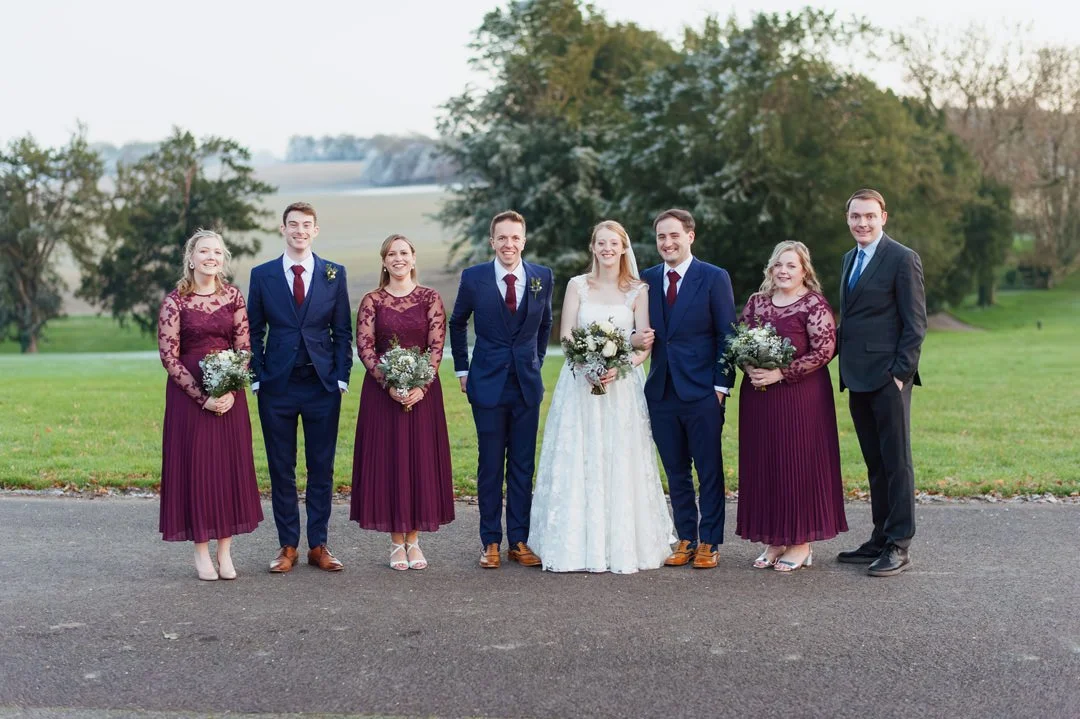 Group of people at a wedding, including a bride in a white dress, groom in a navy suit, and six other guests in formal attire, standing outdoors on a grassy area with trees in the background.