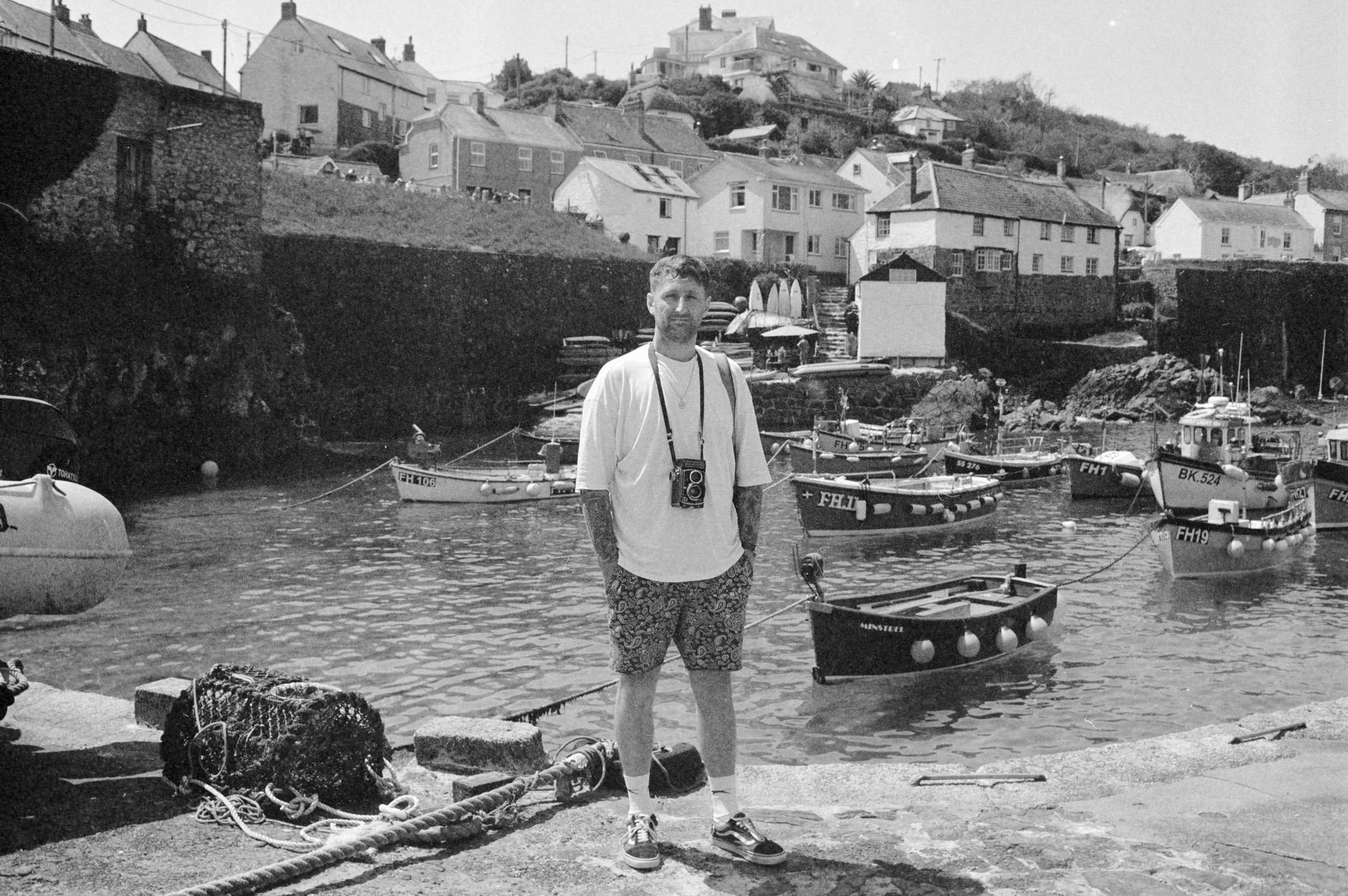 man waring white t shirt stood on harbour wall with boats behind, he has a rolleicord TLR camera around his neck.
