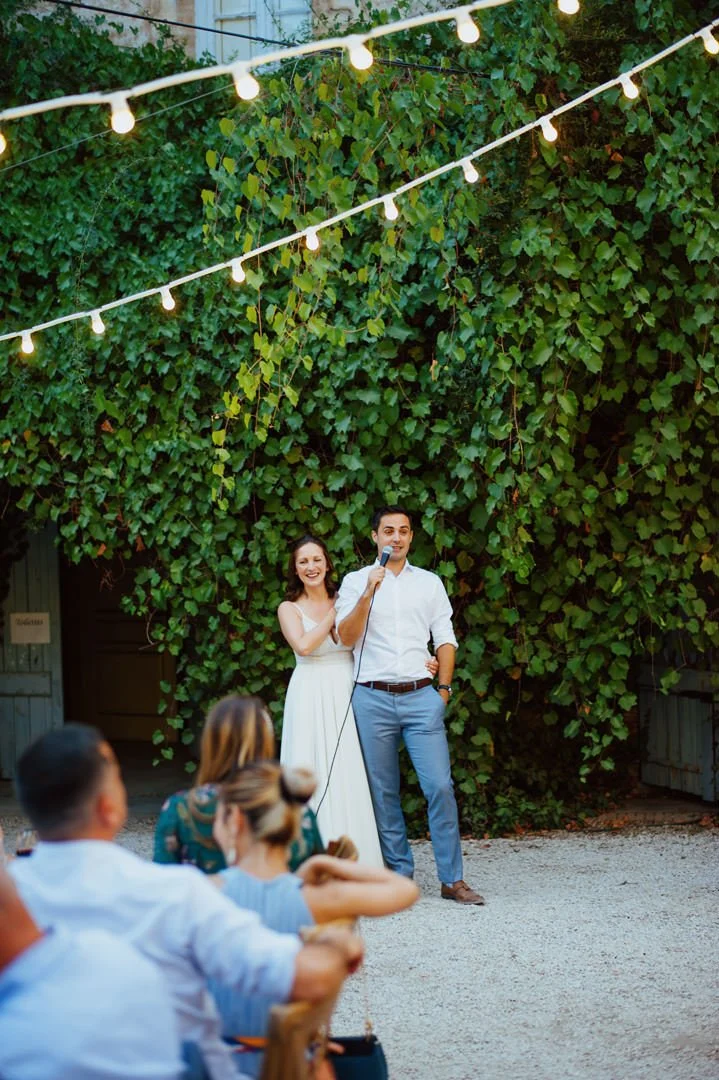 A couple dressed in white stand outdoors under string lights, with the woman smiling and the man speaking into a microphone. Several seated guests are watching the scene.