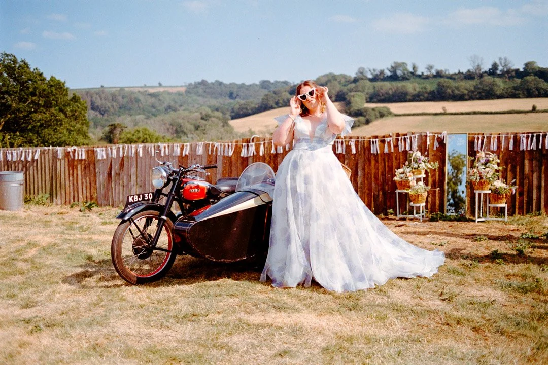 A woman in a long, flowing white dress with puffy sleeves and pink sunglasses standing next to a vintage motorcycle with a sidecar in an outdoor setting with rolling hills and a wooden fence.