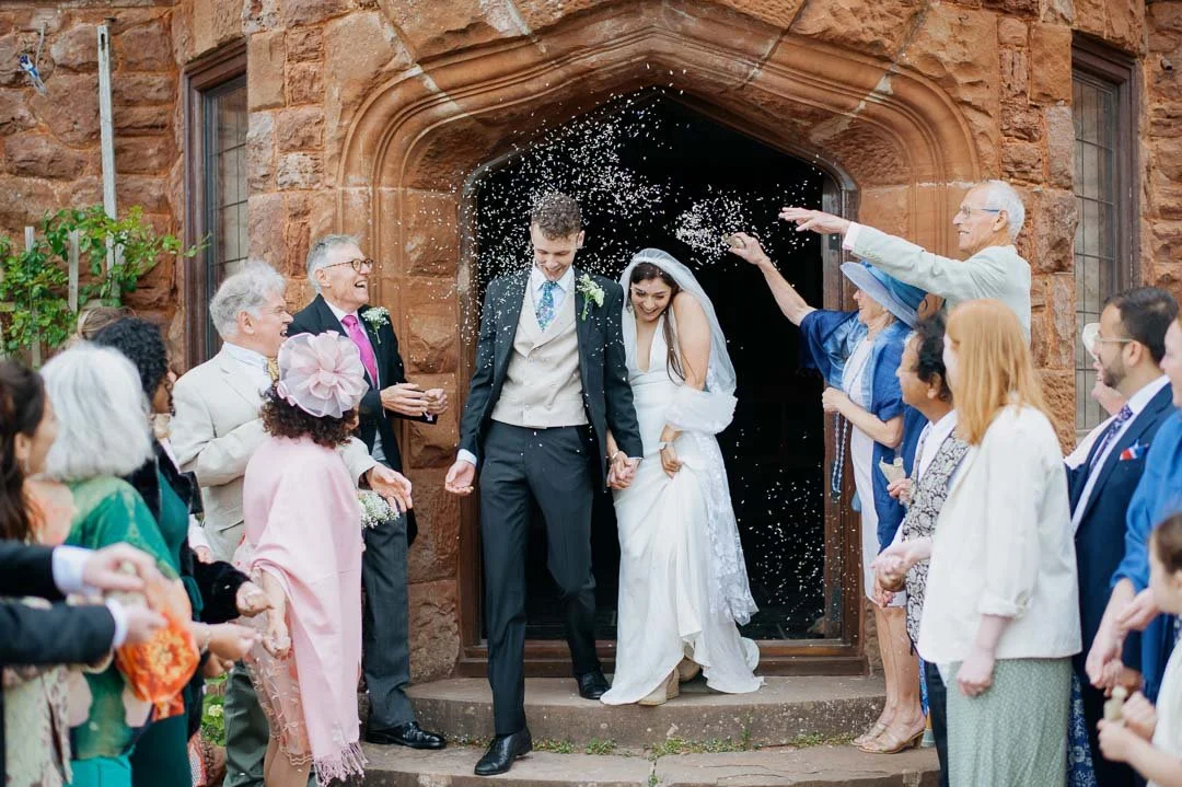 A bride and groom exit a church, holding hands, surrounded by friends and family celebrating with confetti.
