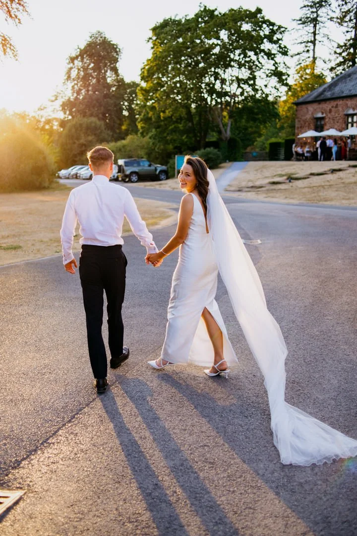 A newlywed couple holding hands and walking away from the camera during sunset, with a large tree and a rustic building in the background.