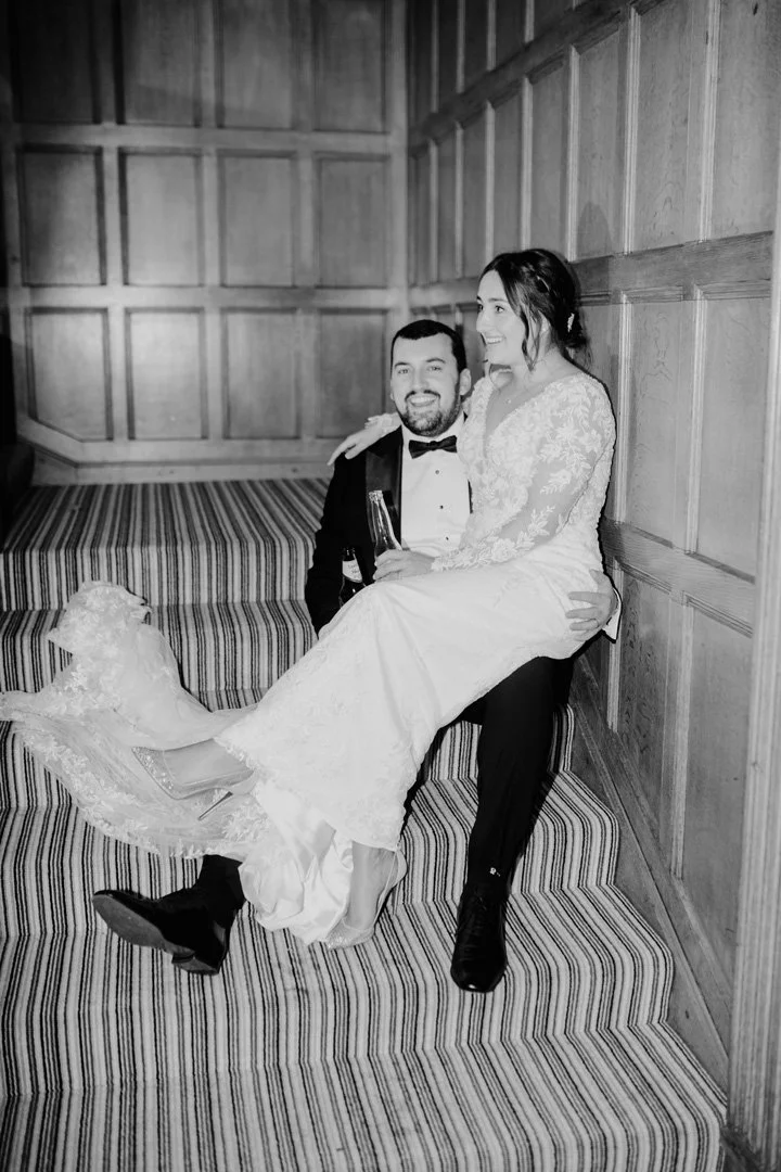A black-and-white photo of a newlywed couple in wedding attire, smiling and sitting on a striped staircase with a wooden-paneled wall in the background. The bride is sitting on the groom's lap, holding a drink, and they appear happy and in love.