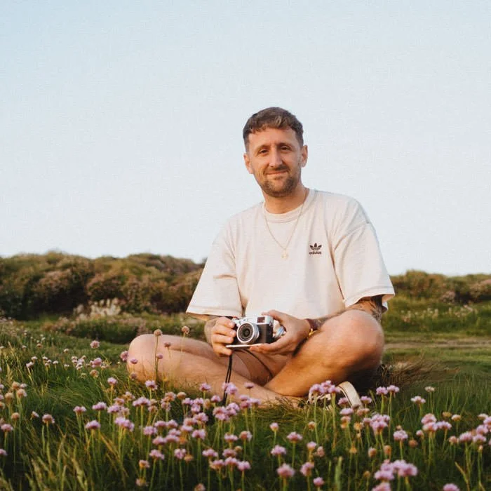 A man sitting cross-legged in a field of pink flowers, holding a camera, with trees in the background during sunset.