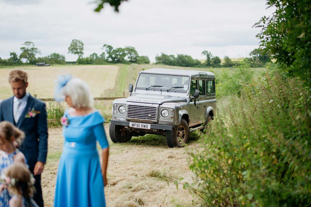 A grey Land Rover Defender parked on a grassy field, with grass and trees in the background, during a daytime outdoor event.