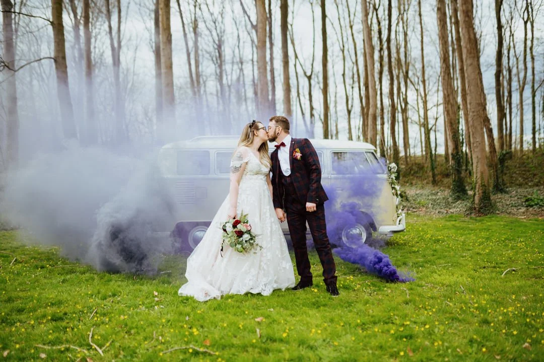 Bride and groom standing on grass field, kissing, with a vintage van in the background emitting purple smoke, in a wooded area.