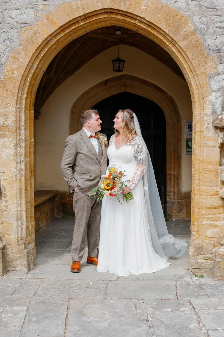A bride and groom standing under an arched stone doorway, smiling at each other. The bride is holding a colorful bouquet, wearing a white lace wedding dress with a veil, and the groom is dressed in a brown tweed suit with an orange bow tie and brown 