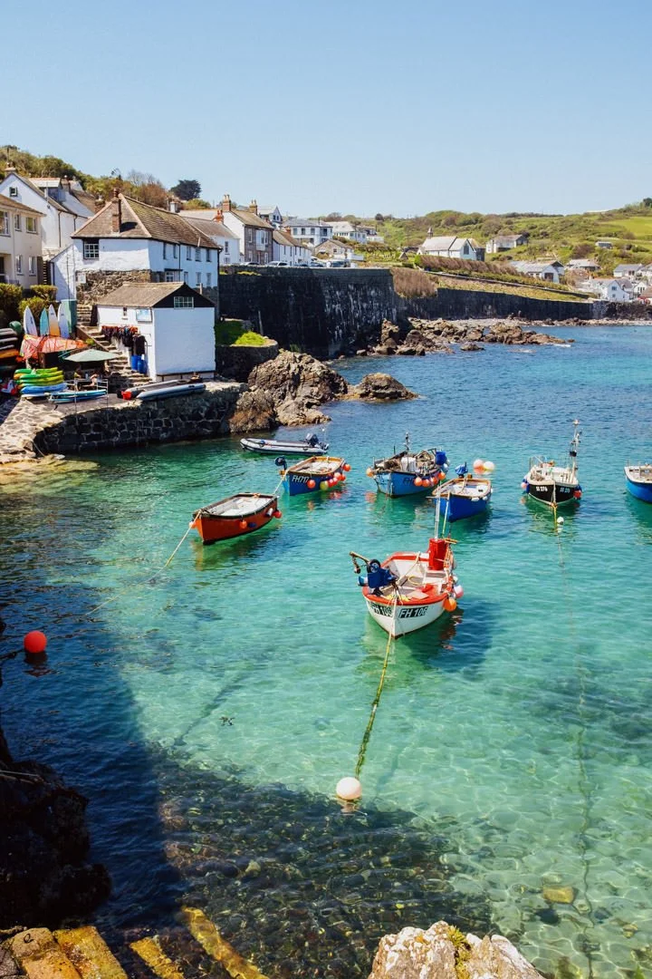 A scenic harbor with small boats floating on clear turquoise water, surrounded by white coastal houses with tiled roofs, some boats anchored, and green hills in the background under a blue sky.