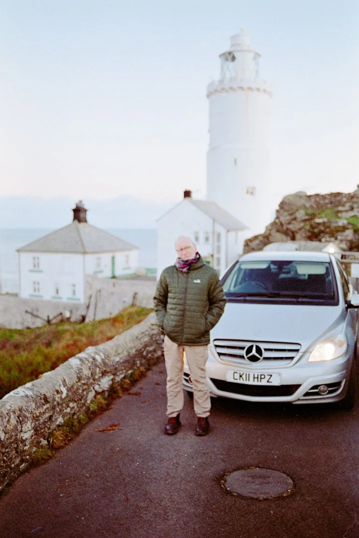 A man standing next to a silver Mercedes-Benz vehicle on a coastal road with a white lighthouse and white houses in the background.