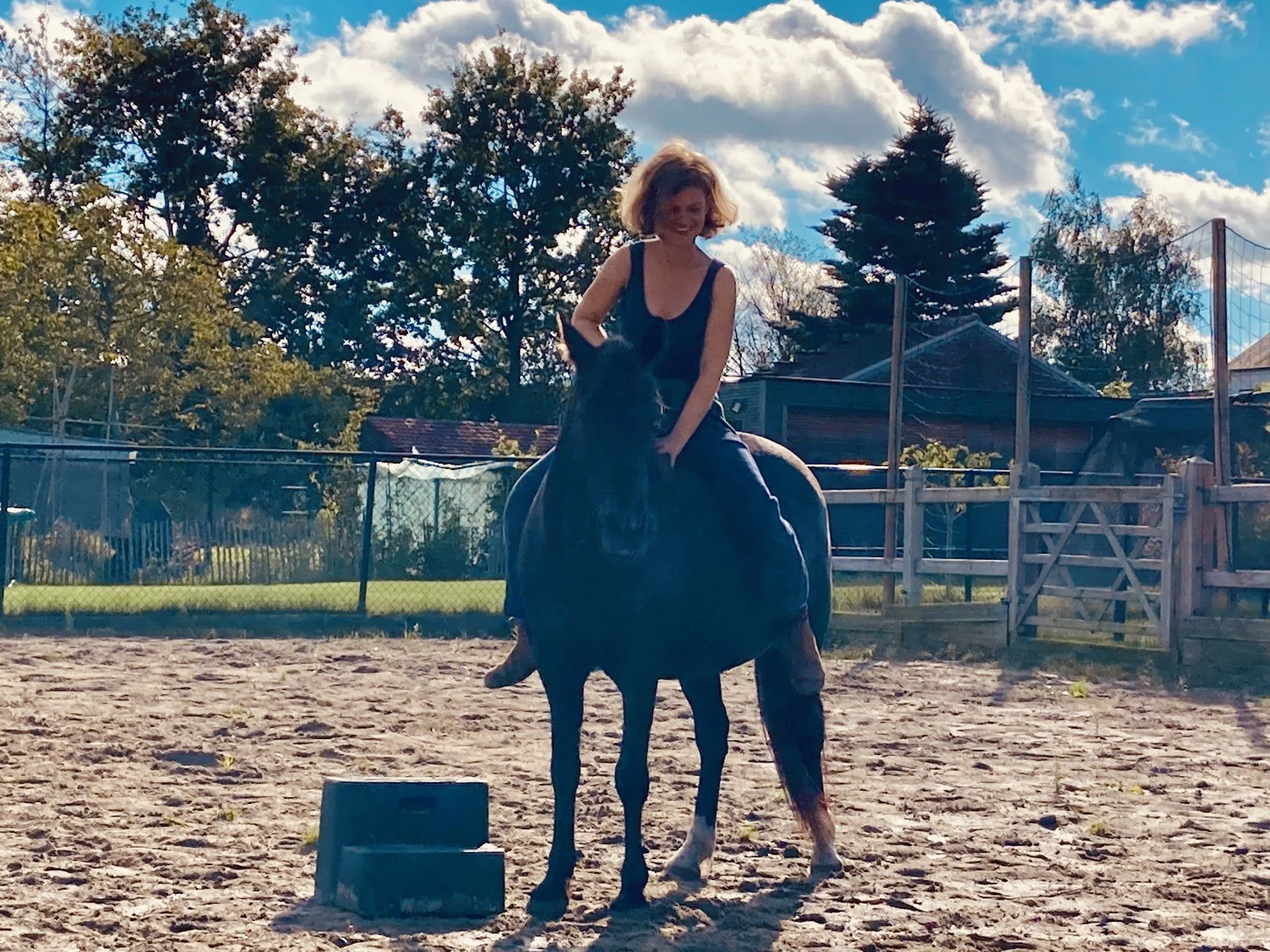 Vrouw zit op een paard in een buitenrijbaan, omringd door een houten omheining met bomen en een blauwe lucht op de achtergrond.