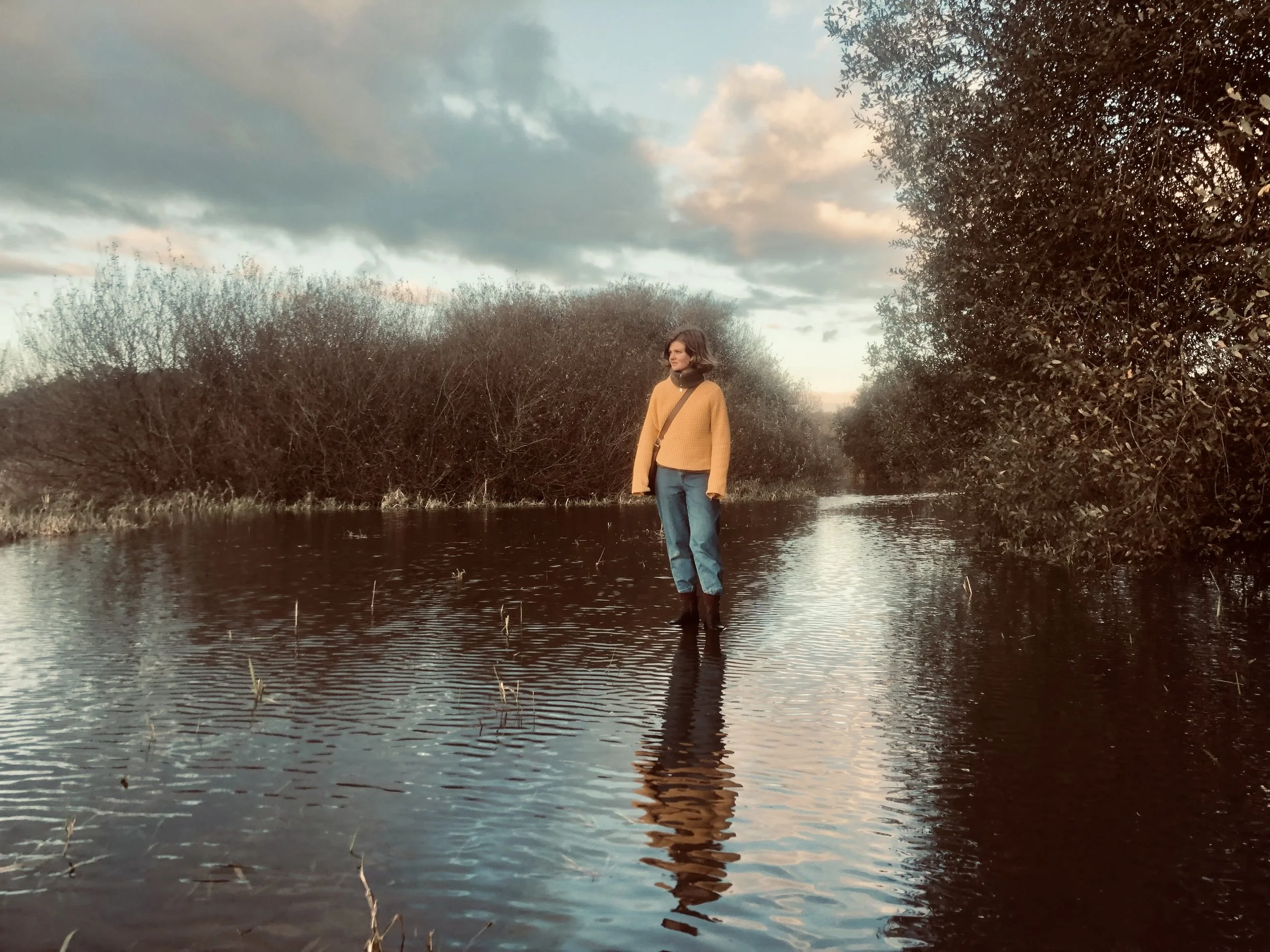 Vrouw in gele trui en blauwe spijkerbroek staat in een waterlijk omgeven door struiken op een bewolkte dag.