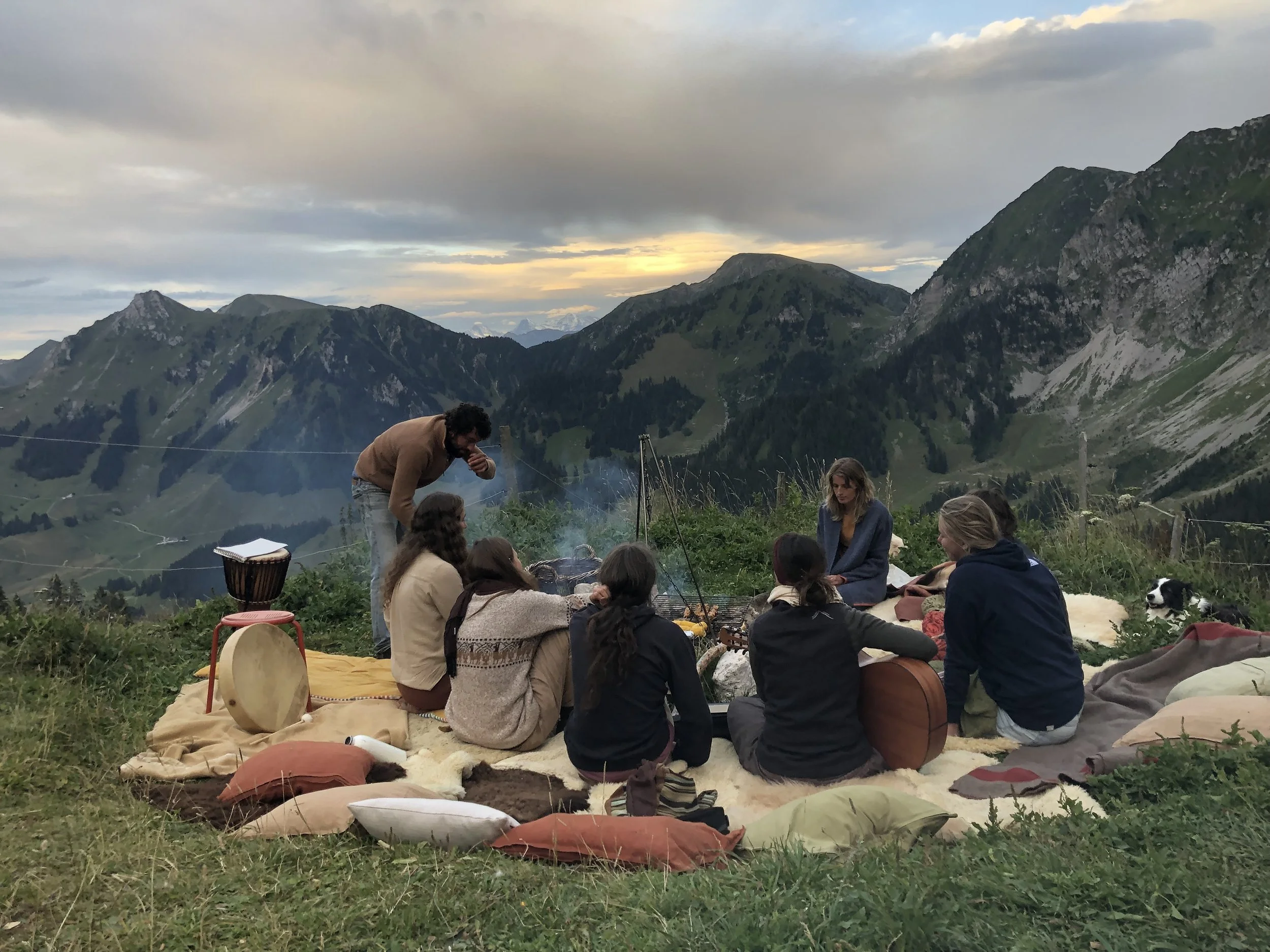 Groep mensen zit op een lappendeken op een berghelling, omringd door kleurrijke kussens, terwijl ze samen muziek maken en genieten van het uitzicht op bergen en een ondergaande zon.