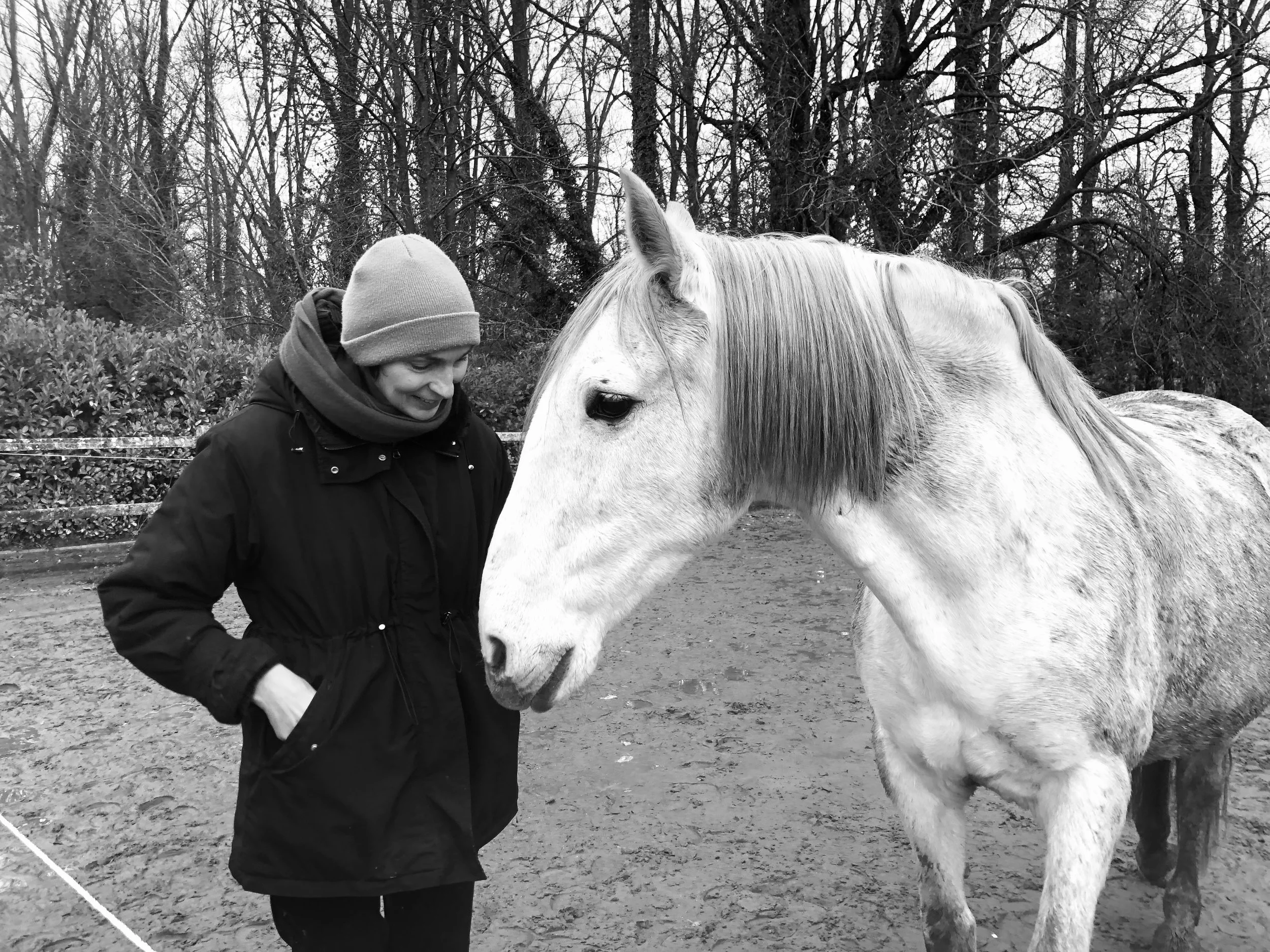 Een persoon in een donkere jas en grijze muts staat naast een witte black-spotted paard op een zandachtig veld, omgeven door bomen zonder bladeren.