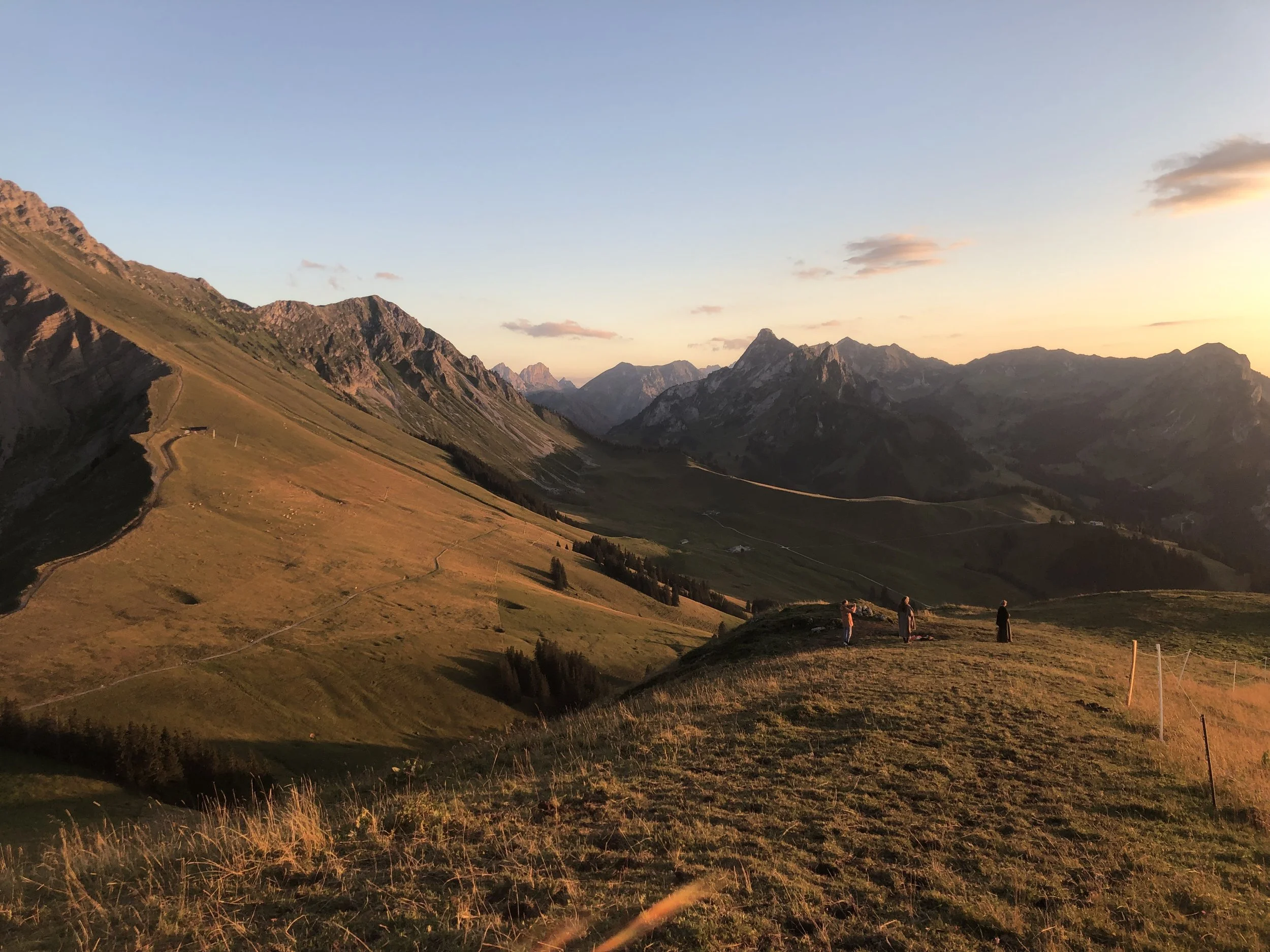 Uitzicht op bergen en vallei tijdens zonsondergang, met drie mensen op een heuvel in de voorgrond.