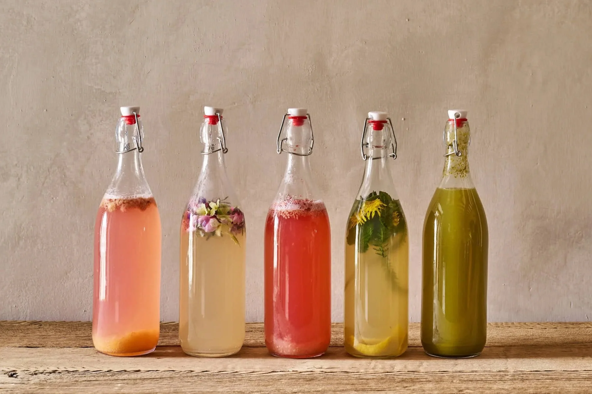 Fünf Glasflaschen mit unterschiedlichen Kräutern und Früchten gefüllt, auf einer Holzoberfläche gegen eine helle Wand