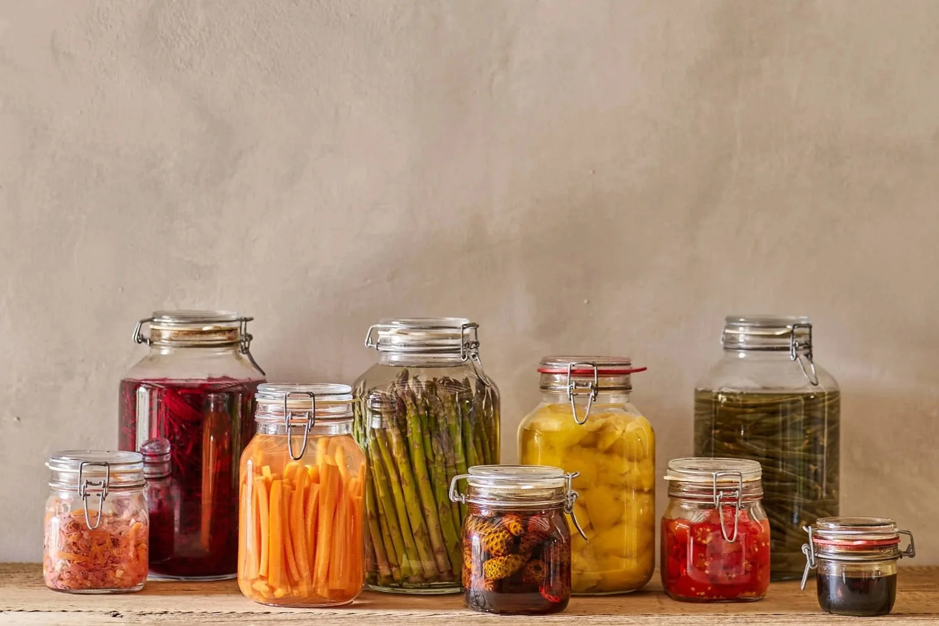 Verschiedene eingemachte Lebensmittel in Gläsern auf einer Holzoberfläche, darunter Rote Beete, Karotten, Spargel, Ananas, grüne Bohnen, Marmelade, Beeren, und schwarze Beeren.