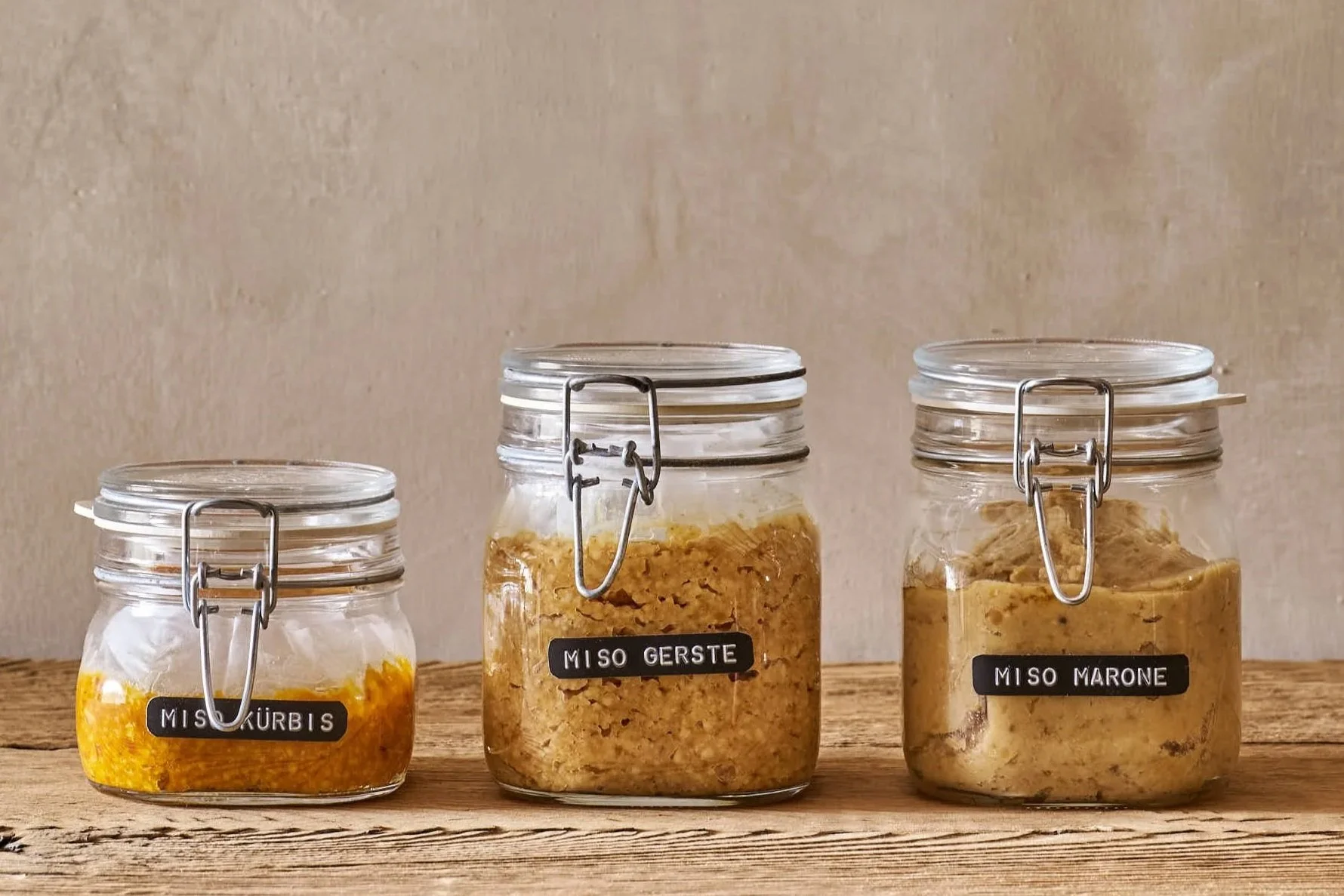 Drei Gläser mit unterschiedlichen Pasten, gekennzeichnet mit schwarzen Labels: 'MISO KÜRbis', 'MISO GERSTE' und 'MISO MARONE', auf einem Holztisch vor einer beigen Wand.