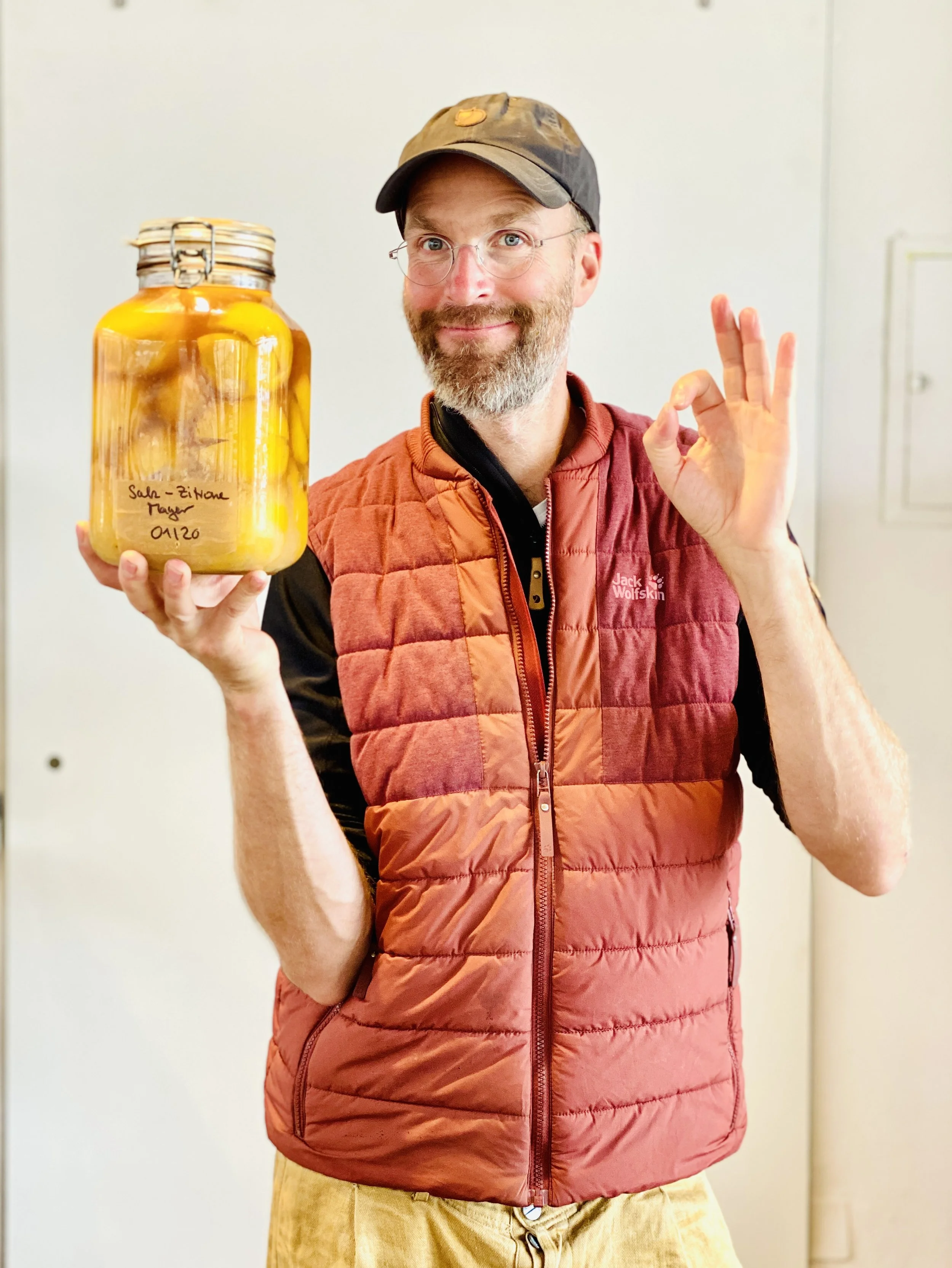 Ein lachender Mann in einer roten Jacke und Mütze hält ein großes Einmachglas mit eingelegtem Gemüse oder Früchten, das mit einem Etikett versehen ist.