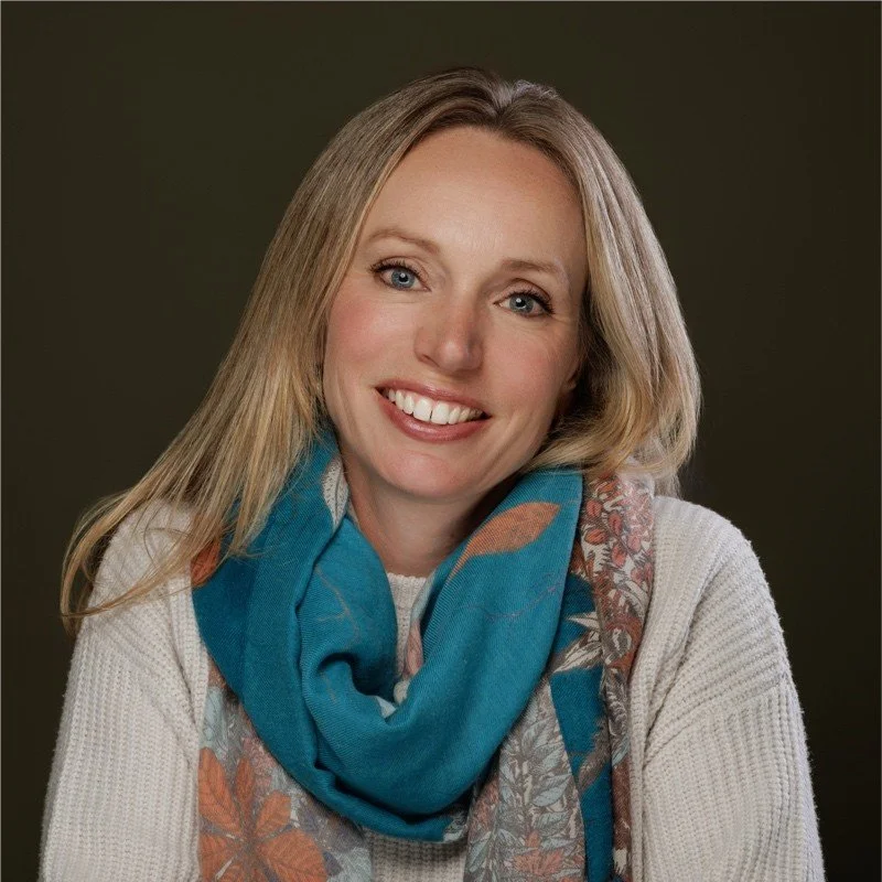 Close-up portrait of a smiling woman with blonde hair, wearing a white sweater and a colorful scarf with blue, orange, and floral patterns against a dark background.