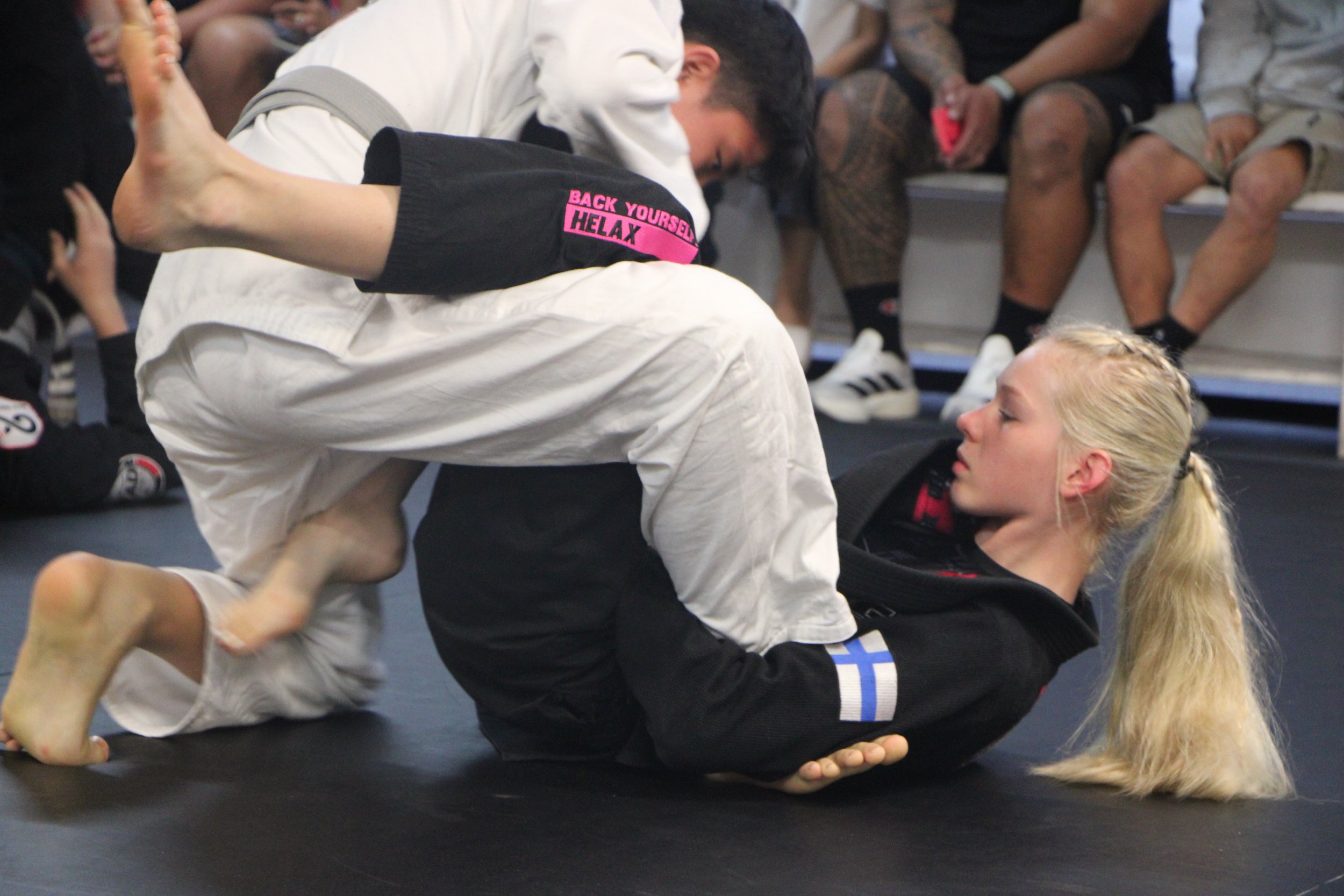 Two kids practicing Brazilian Jiu-Jitsu on a mat, one in a black gi with white stripes and the other in a black gi, with a woman in a gi and a black jacket supervising in the background.