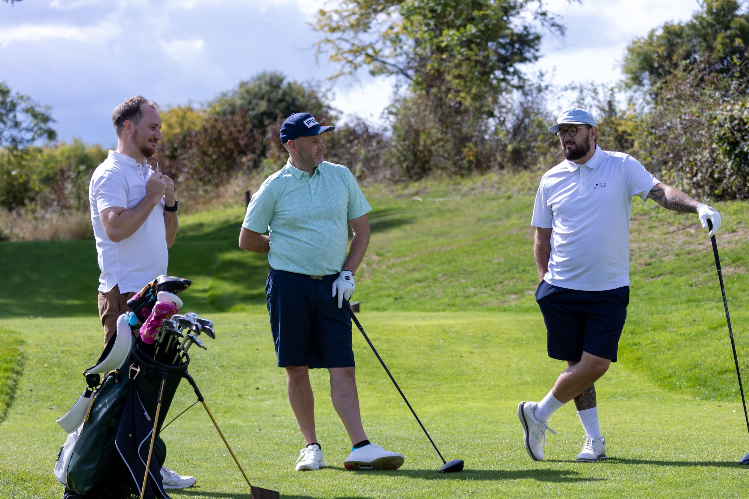 Three men on a golf course, with one man in the middle holding a golf club, another on the right with a golf club resting on the ground, and the third on the left near a golf bag with clubs.