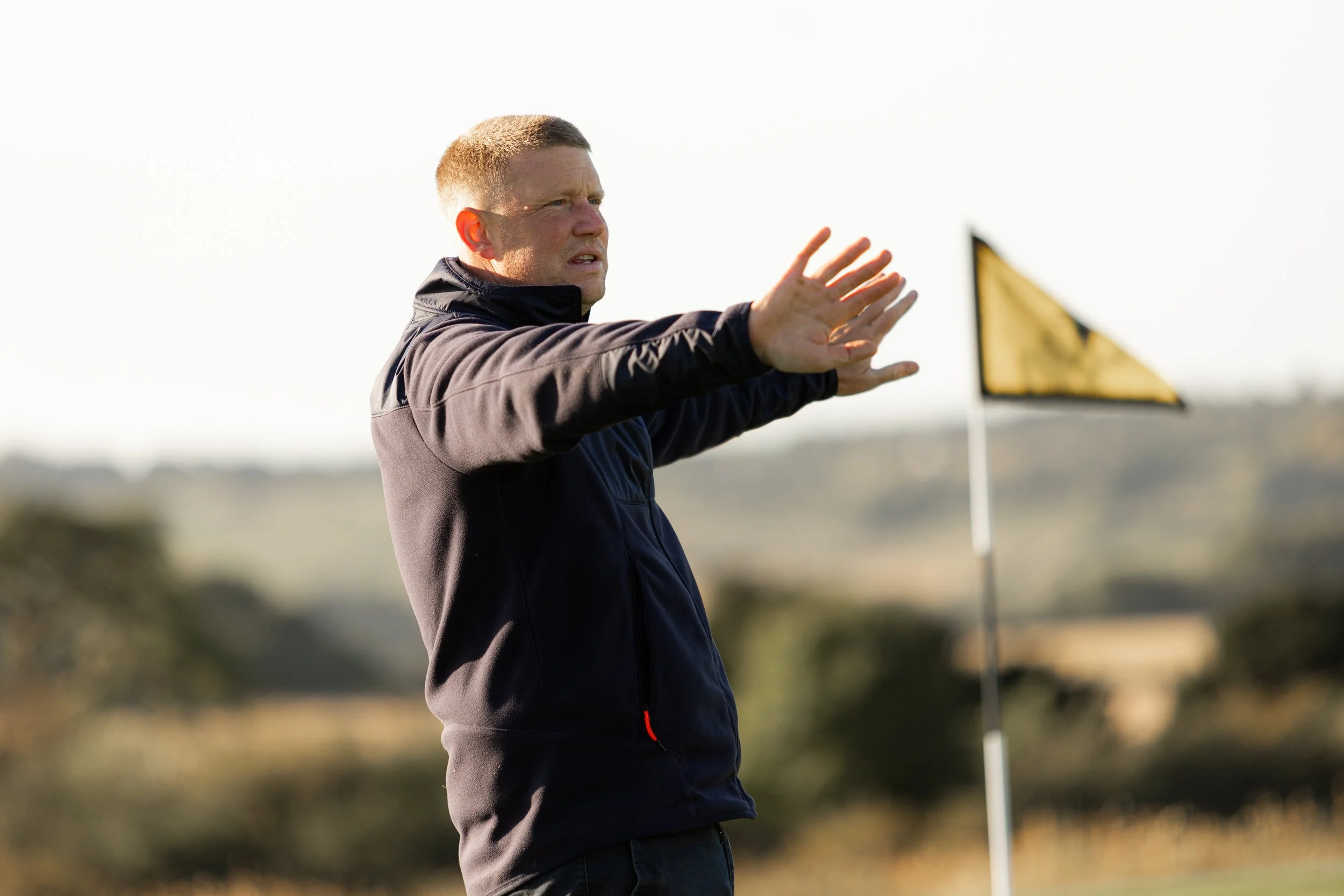 A man was standing near a yellow and black corner flag on a golf course, with his arms extended outward and hands open, in a gesture of signaling or giving instructions, with a blurry background of fields and trees.