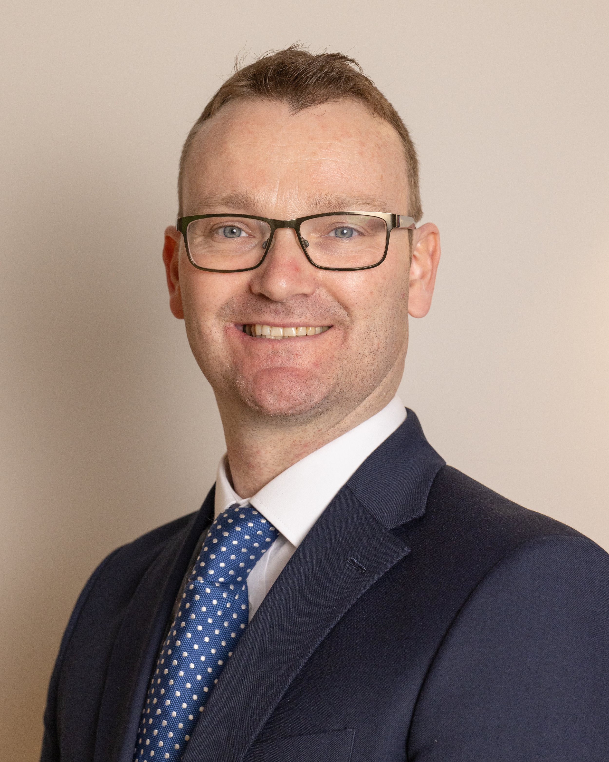 Professional headshot of a man in a dark suit, white shirt, blue polka-dot tie, wearing glasses, smiling, short brown hair, against a plain background.